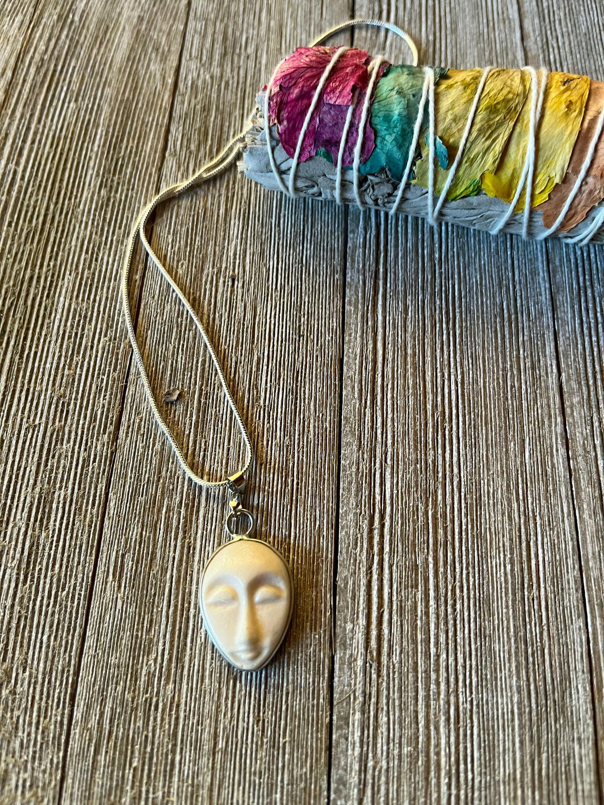 The Serene Carved Bone Face Necklace—Tranquil Talisman for Calm &amp; Clarity—rests beside dried sage and flower petals, offering a unique spiritual accessory to inspire mindfulness and peace.