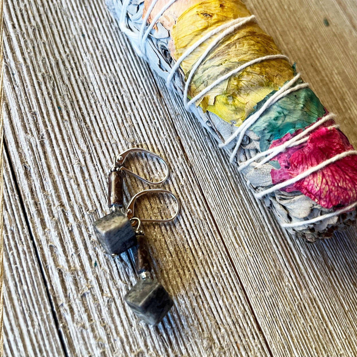 The Sacred Geometry Dark Jasper Cube Earrings—Sterling Silver Artisan Jewelry—feature geometric dark jasper cubes on silver hooks, displayed on wood beside a colorful sage smudge stick wrapped in white string.