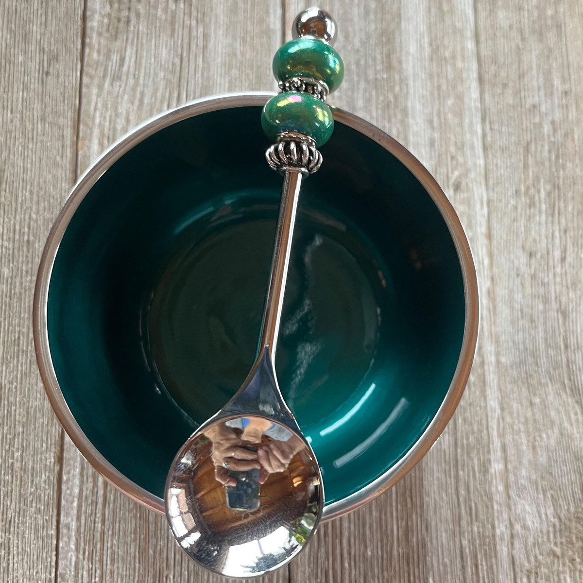 The Festive Green Beaded Spoon with Stainless Steel Bowl Set, a perfect condiment holder for parties, sits on a wooden surface. The spoon’s shiny handle features green beads and reflects hands holding a camera.