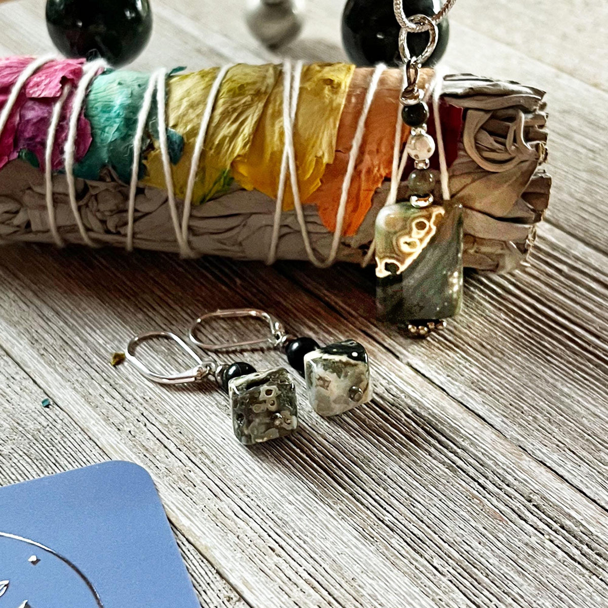 The Ocean Jasper Jewelry Set—Zen minimalist necklace and earrings crafted from rare natural stones—rests beside colorful smudge sticks on a wooden surface.