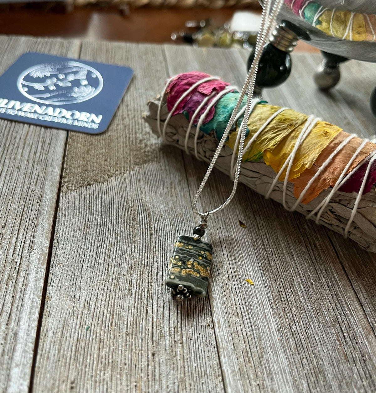 The Ocean Jasper Winter Seas Pendant, a minimalist silver necklace with a rectangular green and gold stone, rests on rustic wood next to a vibrant sage bundle and a business card.