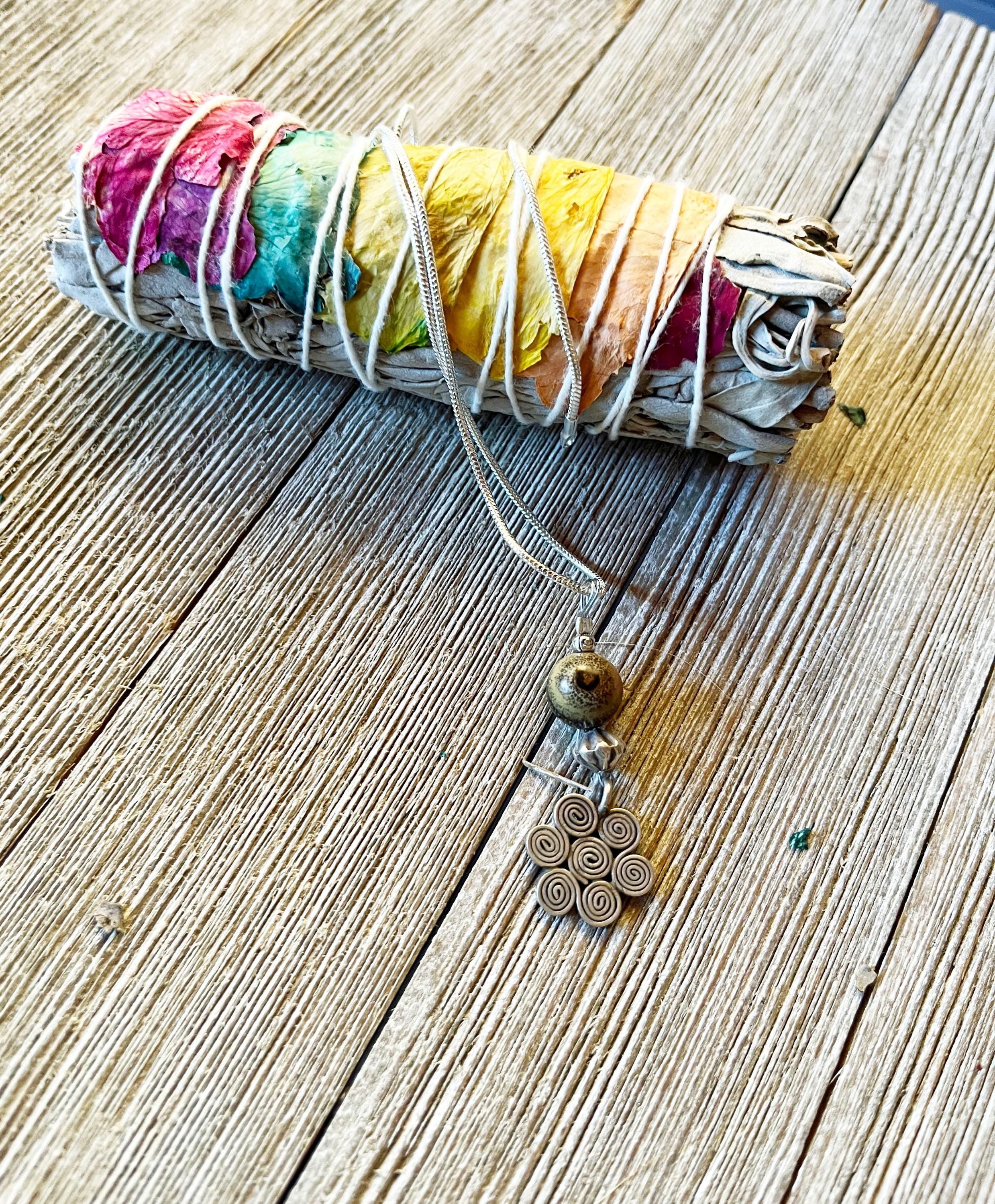 A bundle of sage with dried rose petals sits beside the Swirling Winds Pendant—a minimalist winter necklace featuring a rare natural stone and spiral pattern.
