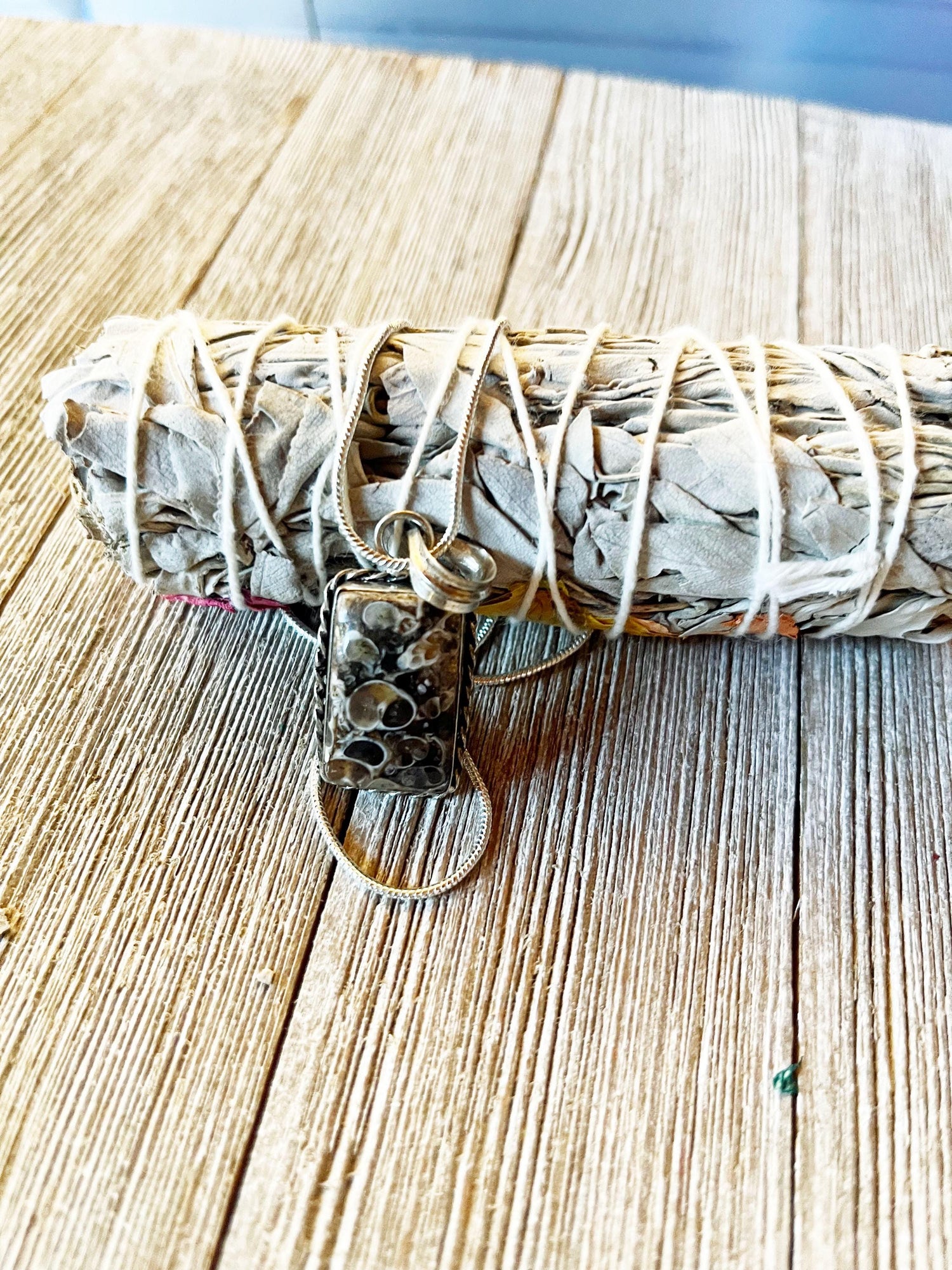 A bundle of dried sage lies on wood, adorned with a keychain holding the Fossil Jasper Pendant – a gift as old as time for the winter season – and two metal rings, bringing grounding energy to your space.