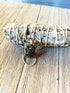 A bundle of dried sage lies on wood, adorned with a keychain holding the Fossil Jasper Pendant – a gift as old as time for the winter season – and two metal rings, bringing grounding energy to your space.