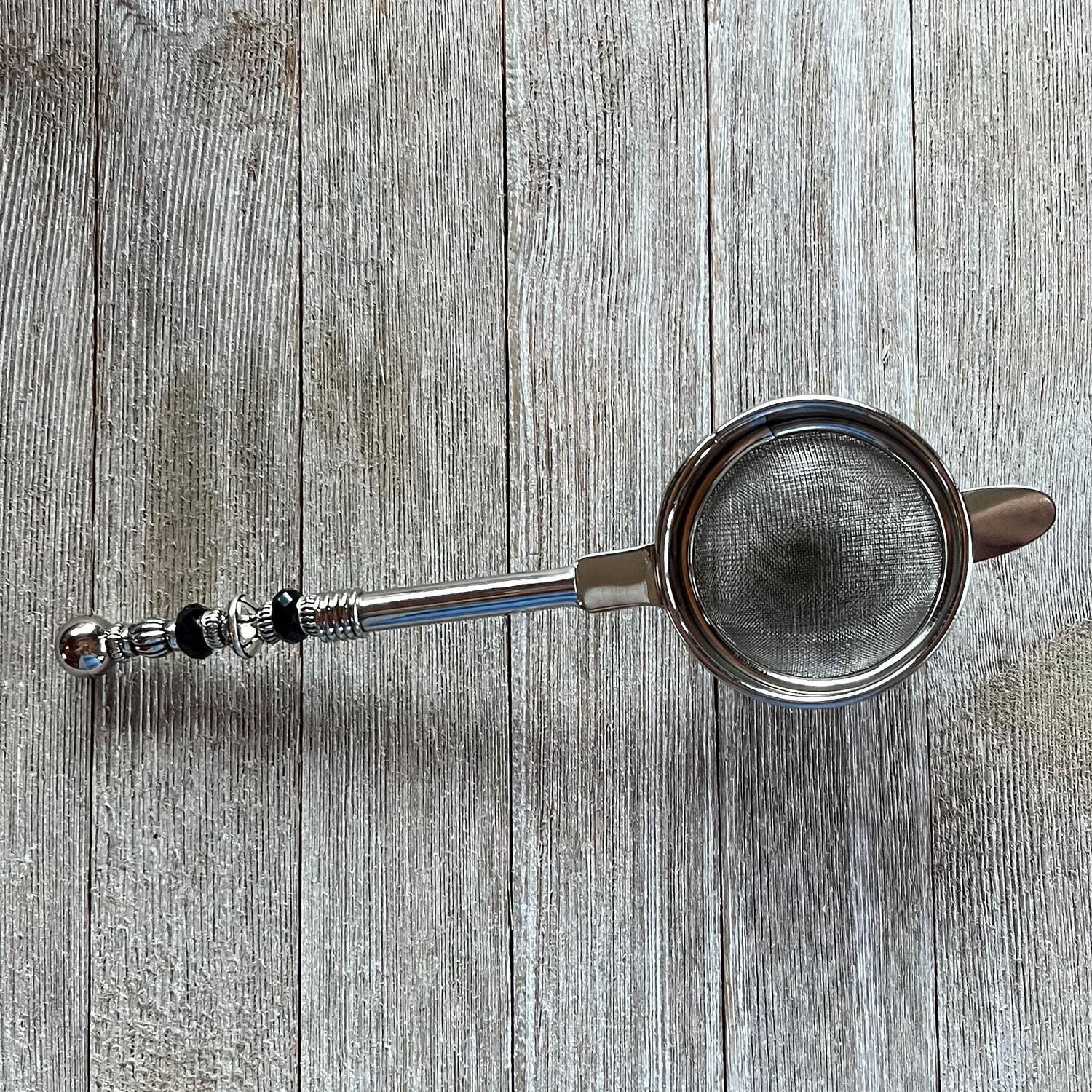 The Beaded Tea Strainer – Handcrafted Customizable Handle (9” Length), adorned with black and silver accents, rests elegantly on a light wooden surface—an ideal gift for tea lovers.