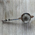 The Beaded Tea Strainer – Handcrafted Customizable Handle (9” Length), adorned with black and silver accents, rests elegantly on a light wooden surface—an ideal gift for tea lovers.