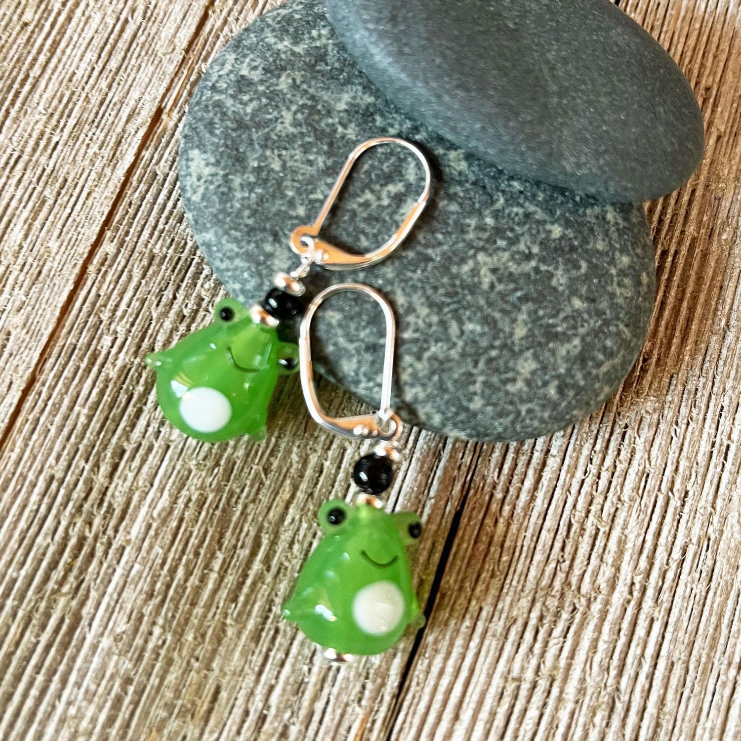 Handcrafted Glass Frog Earrings: Animal Lover Jewelry Gift featuring silver hooks and frog charms, elegantly displayed on stacked gray stones atop rustic wood—a delightful accessory for any animal jewelry enthusiast.
