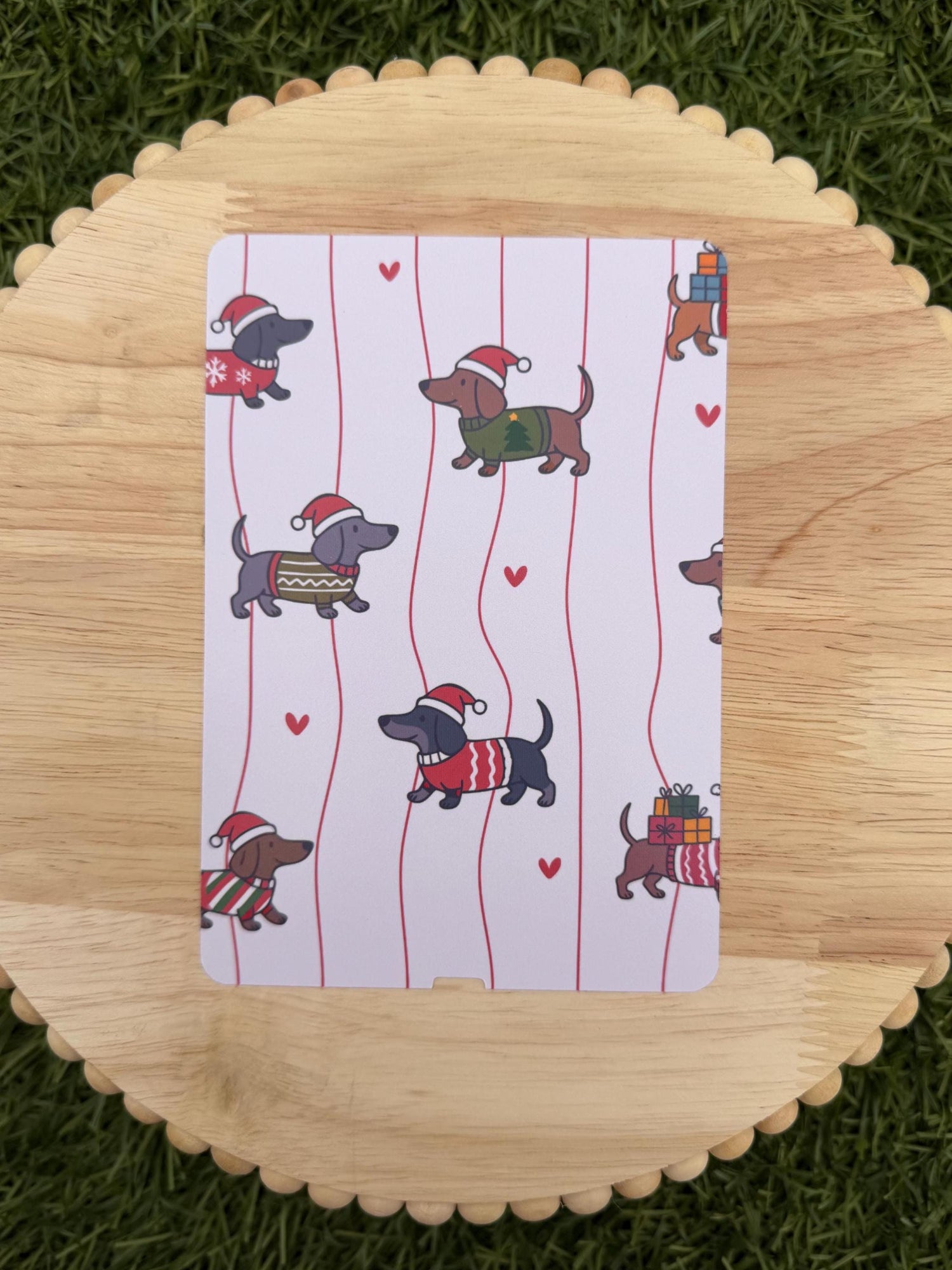 May include: A white card with a repeating pattern of dachshunds wearing Santa hats and sweaters, set against red vertical stripes and small red hearts. The dachshunds are in various colors and poses.