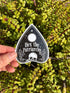 May include: A black and white sticker in the shape of a planchette, with the words "Hex the Patriarchy" in white script. The sticker features a skull, moon, and stars design. The sticker is held in front of a background of green leaves.
