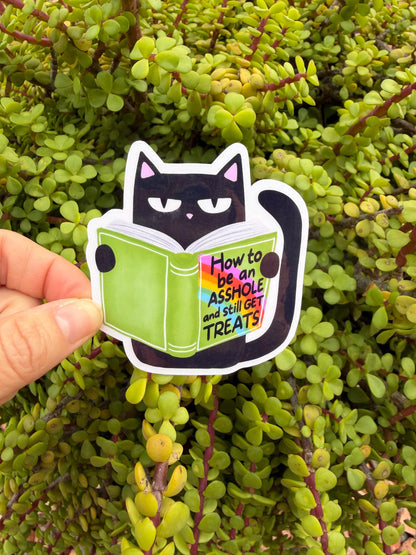 May include: A black cat sticker reading a green book with the text &amp;quot;How to be an ASSHOLE and still GET TREATS&amp;quot;. The cat has pink inner ears and is set against a background of green leaves.