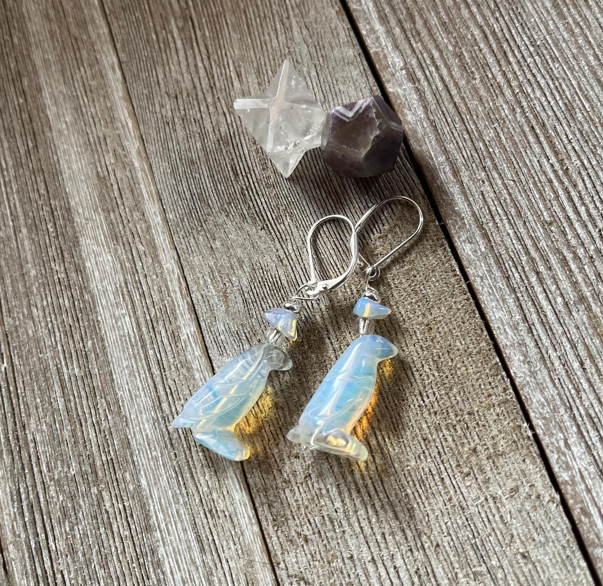 Handcrafted Opalite Penguin Earrings with sterling silver lever backs rest on rustic wooden planks, accompanied by a clear star-shaped crystal and a faceted purple stone.