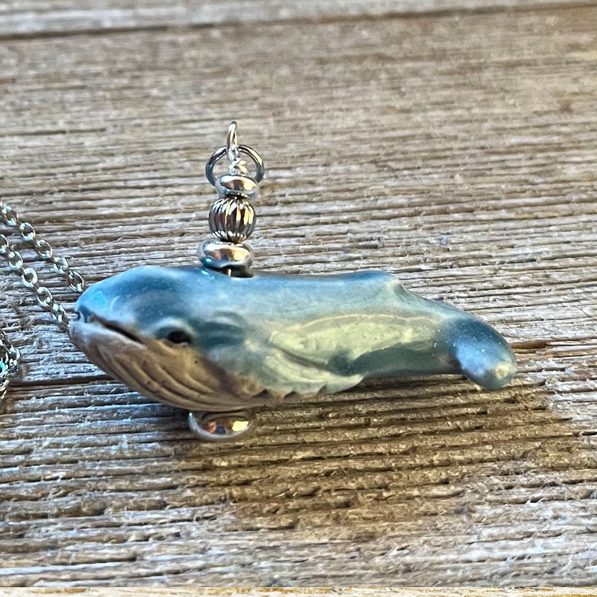 Showcasing the Blue Whale Necklace—an ocean-themed jewelry piece with silver beads and chain, shaped like a blue whale and displayed on textured wood. Add matching earrings if you like! Perfect for whale lovers.