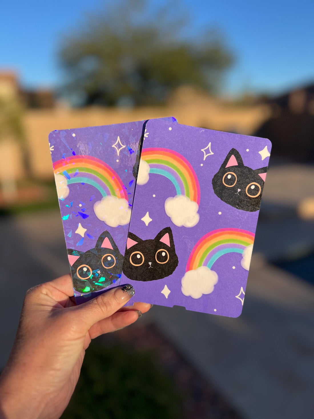 May include: Two purple notebooks with a repeating pattern of black cat faces, rainbows, and stars. One notebook has a holographic foil effect. The notebooks are held up against a blurred outdoor background.