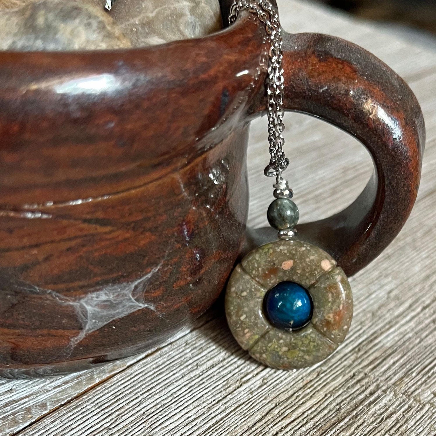 The One of a Kind Jasper Mood Pendant, featuring a color-changing bead on an 18” silver chain, hangs from the handle of a brown ceramic mug resting on a wooden surface.