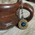The One of a Kind Jasper Mood Pendant, featuring a color-changing bead on an 18” silver chain, hangs from the handle of a brown ceramic mug resting on a wooden surface.