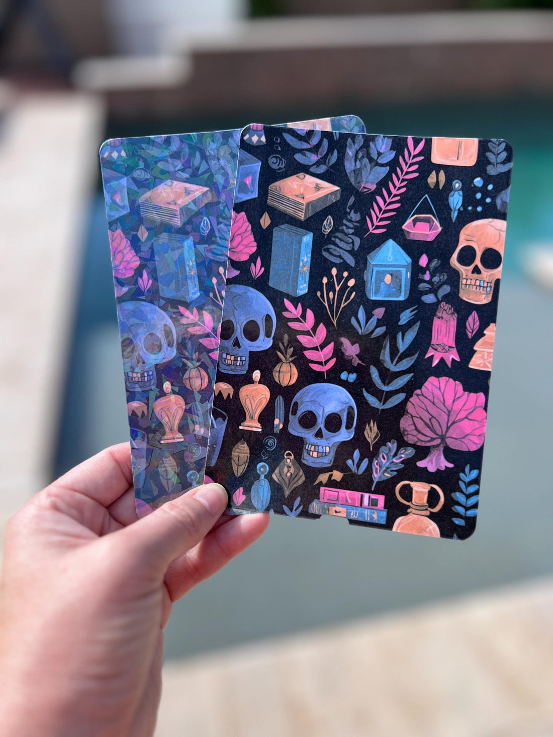 May include: Two decorative cards with a dark background, featuring skulls, books, and botanical elements in shades of blue, pink, and orange. The cards have a mystical and gothic theme, with a detailed and artistic design.
