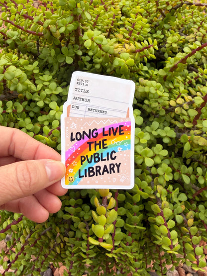 May include: A sticker with a library card design and the text &amp;quot;LONG LIVE THE PUBLIC LIBRARY.&amp;quot; The sticker features a rainbow, stars, and hearts. The background is a green plant.