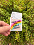 May include: A sticker with a library card design and the text "LONG LIVE THE PUBLIC LIBRARY." The sticker features a rainbow, stars, and hearts. The background is a green plant.