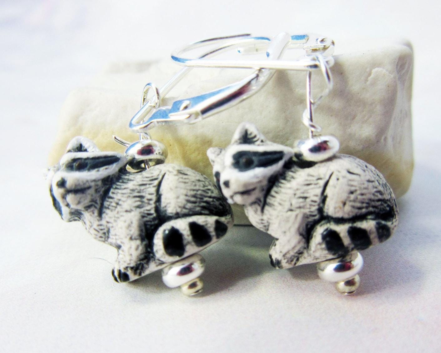 A pair of Raccoon Earrings featuring handpainted garbage panda charms with silver lever backs—perfect as a nature lover gift—displayed in front of a pale stone background.