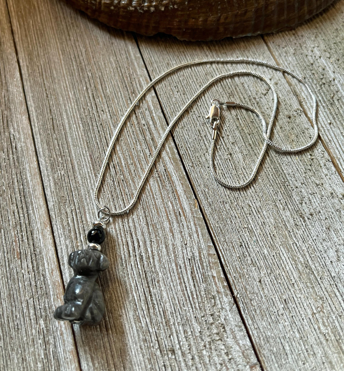 The Carved Obsidian Dog Necklace - Color Change Bead, Pet Memorial features a small dog pendant and a color-changing bead, displayed on a rustic wooden surface.