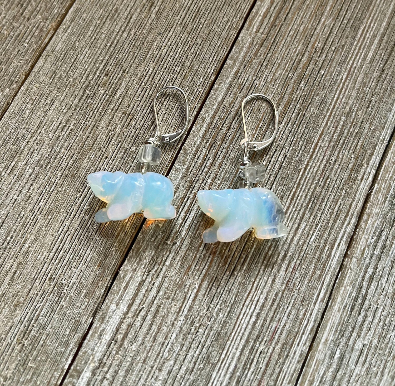 Handcrafted Stone White Bear Earrings shimmer like the northern lights with iridescent bear charms and clear beads on sterling silver lever backs, beautifully displayed on a rustic wooden surface.