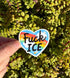 May include: A heart-shaped sticker with the words "Fuck ICE" in black, over a rainbow and blue sky background. The sticker has smiley faces and small white flowers. The sticker is held in front of a background of green leaves.