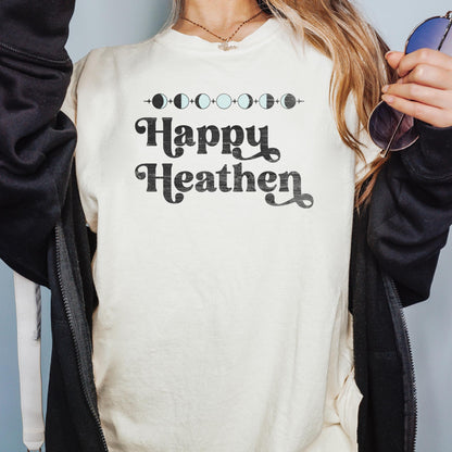 May include: Cream-colored t-shirt with the text &amp;quot;Happy Heathen&amp;quot; in a black, vintage-style font. Above the text is a line of moon phase graphics. The shirt is a casual, everyday wear item.