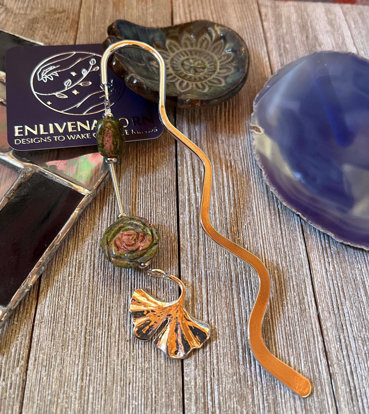The Unakite Flower Metal Bookmark: Zen Ginko Leaf Design features green and rose-like beads on a wavy metal shape, displayed on rustic wood amid stained glass and ceramic dishes—an ideal reader gift.