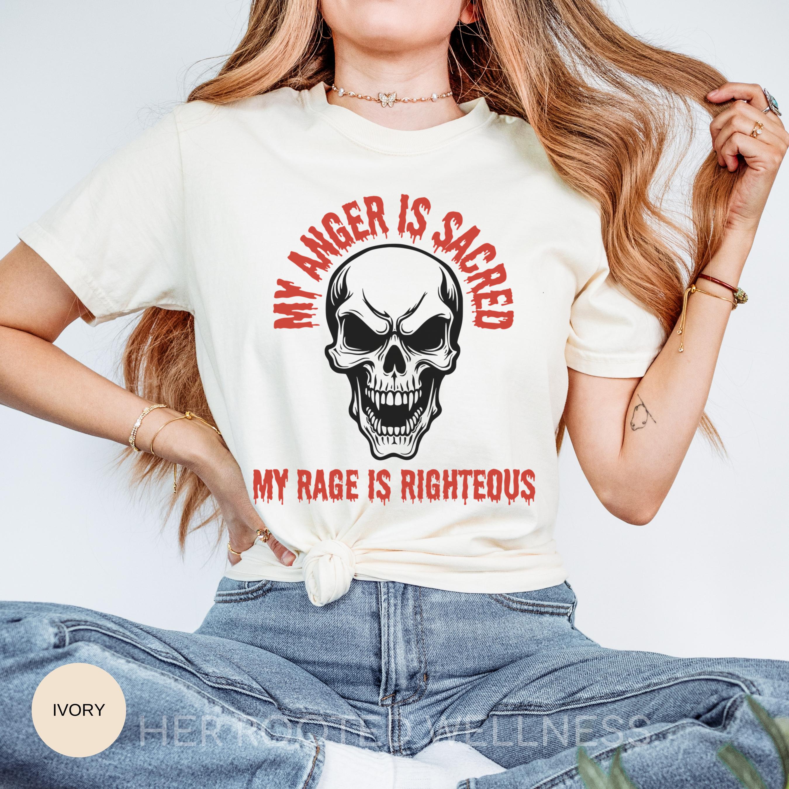 May include: Ivory-colored t-shirt with a skull graphic and the text &amp;quot;MY ANGER IS SACRED&amp;quot; and &amp;quot;MY RAGE IS RIGHTEOUS&amp;quot; in red. The skull has a menacing expression. The shirt is a casual wear item.