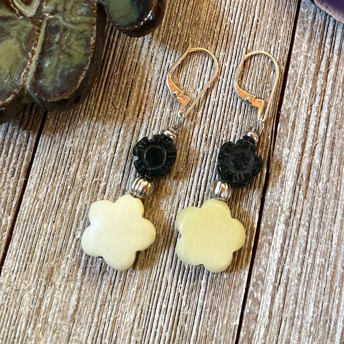Spring Flower Earrings in carved stones, featuring cream flower-shaped beads, black carved beads, and silver spacers, perfect to complete your wardrobe and add cheerful style to daily wear. Displayed on a wooden surface by a ceramic object.