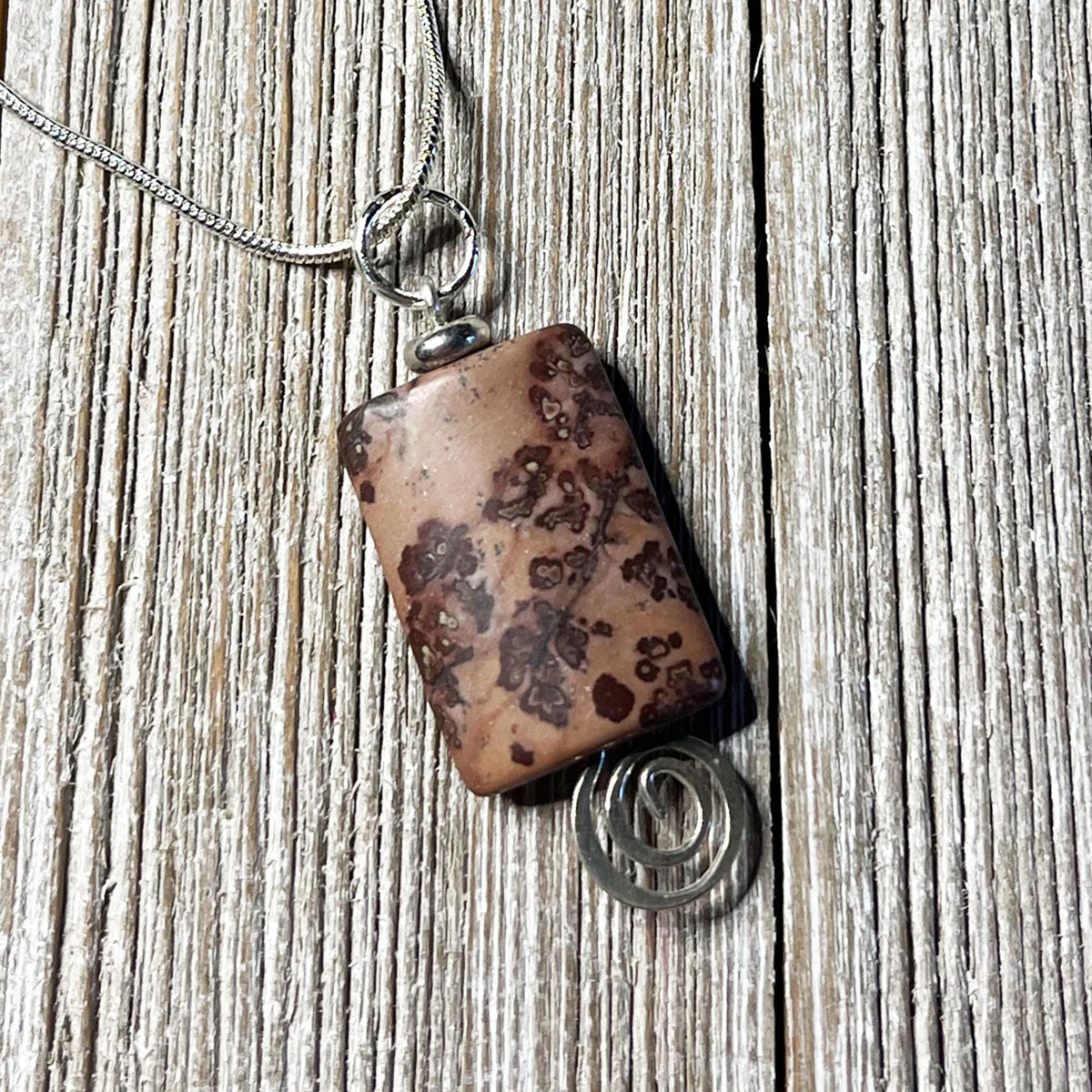 The Crazy Horse Jasper Necklace: Minimalist Zen Pendant, handcrafted in the USA, features a brown stone with dark patterns in a silver wire setting with a spiral accent and hangs from a silver chain on rustic wood.