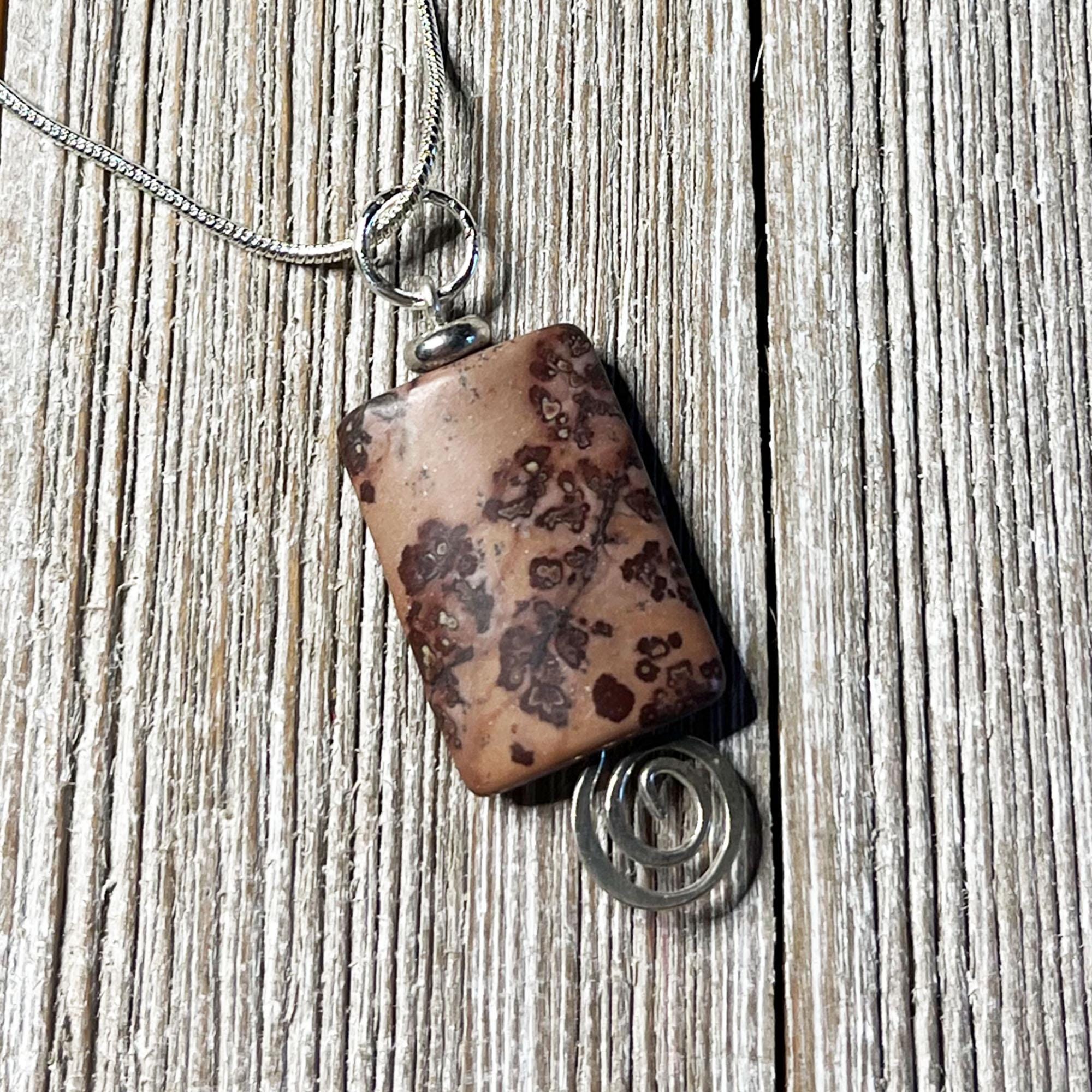 The Crazy Horse Jasper Necklace: Minimalist Zen Pendant, handcrafted in the USA, features a brown stone with dark patterns in a silver wire setting with a spiral accent and hangs from a silver chain on rustic wood.