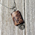 The Crazy Horse Jasper Necklace: Minimalist Zen Pendant, handcrafted in the USA, features a brown stone with dark patterns in a silver wire setting with a spiral accent and hangs from a silver chain on rustic wood.