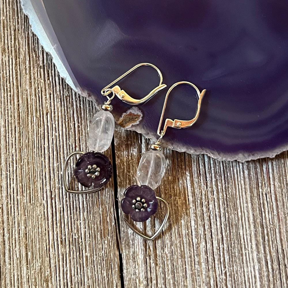 Violet Flower Earrings: Spring Flower Jewelry featuring silver hooks, clear oval beads, dark purple flower beads, and silver heart charms—a unique handmade gift or spiritual gift for wisdom. Shown on wood near a purple agate slice.