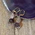 Violet Flower Earrings: Spring Flower Jewelry featuring silver hooks, clear oval beads, dark purple flower beads, and silver heart charms—a unique handmade gift or spiritual gift for wisdom. Shown on wood near a purple agate slice.