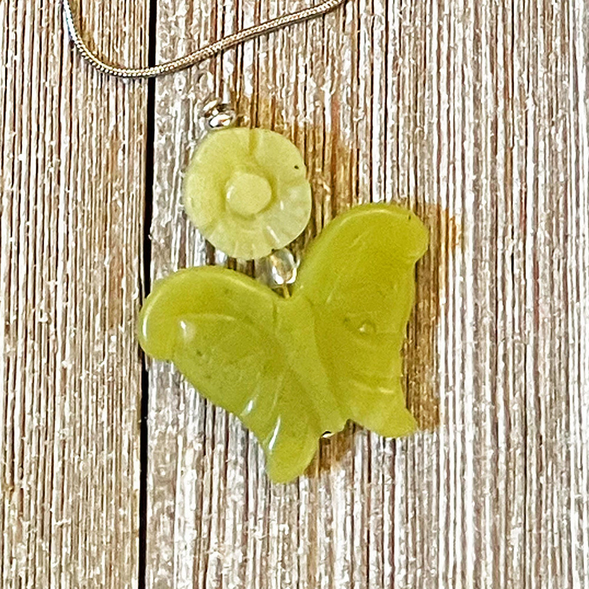 The Carved Jadeite Butterfly Pendant with Flower – Spring Nature Jewelry features a green butterfly pendant on a silver chain, beautifully showcased on textured wood.