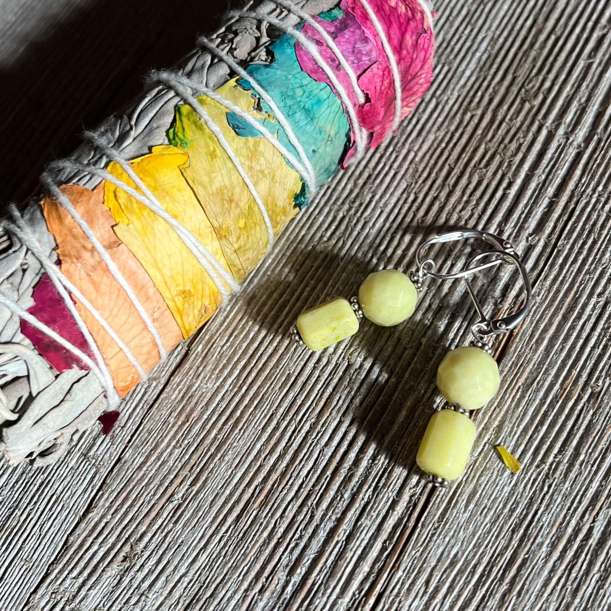 A bundle of dried sage with colorful threads rests beside Spring Earrings featuring green howlite stones—perfect for brightening up your wardrobe—on a textured wooden surface.