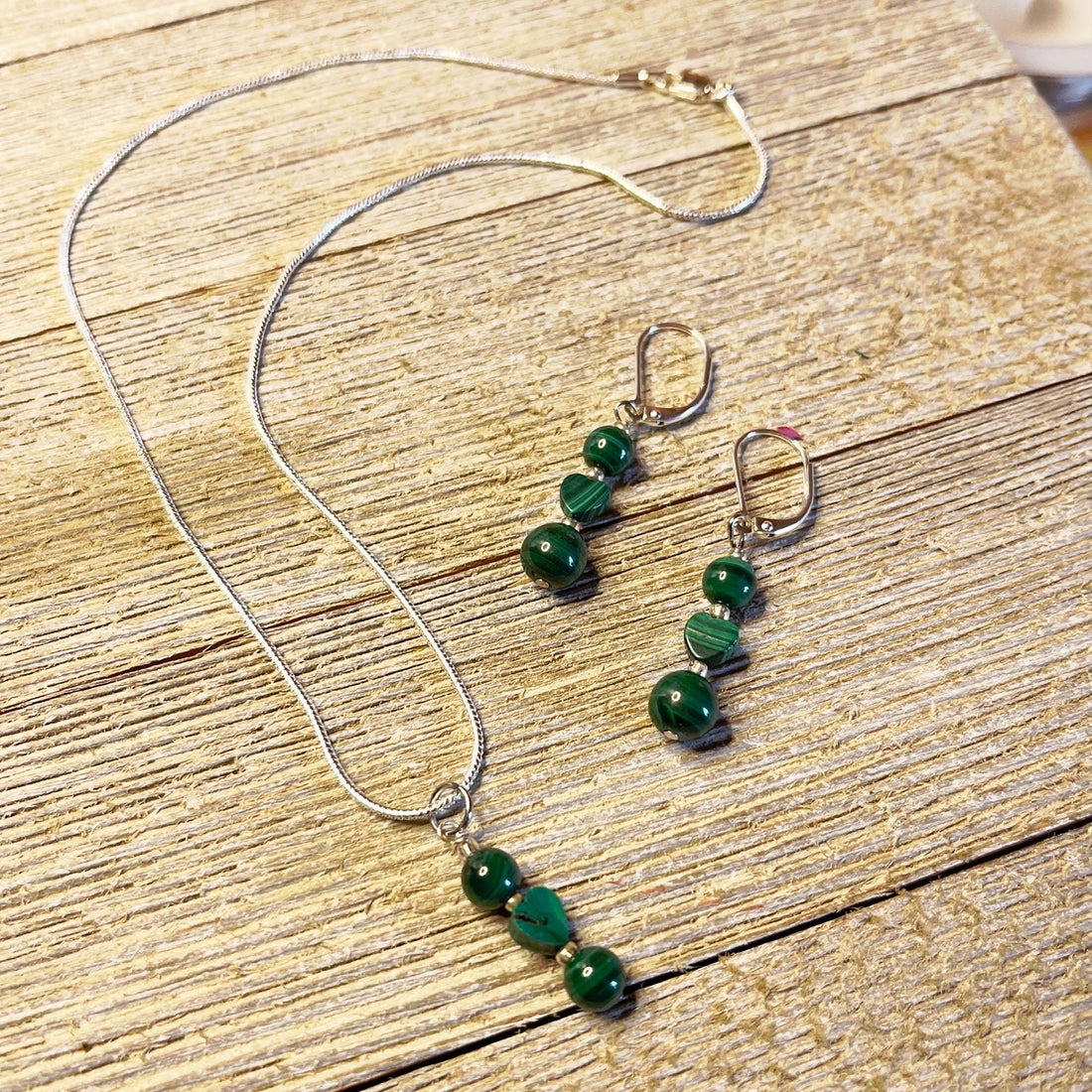 The Green Malachite Heart Jewelry Set features a Spring Dangle Necklace with three green striped beads in a row and matching earrings with two beads each—an elegant handcrafted set displayed on light wood, perfect for any occasion.