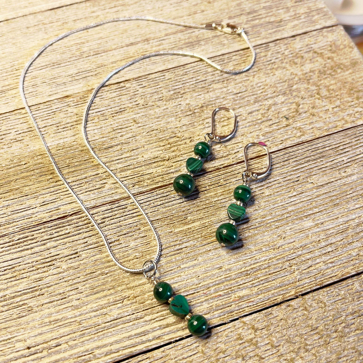 The Green Malachite Heart Jewelry Set features a Spring Dangle Necklace with three green striped beads in a row and matching earrings with two beads each—an elegant handcrafted set displayed on light wood, perfect for any occasion.