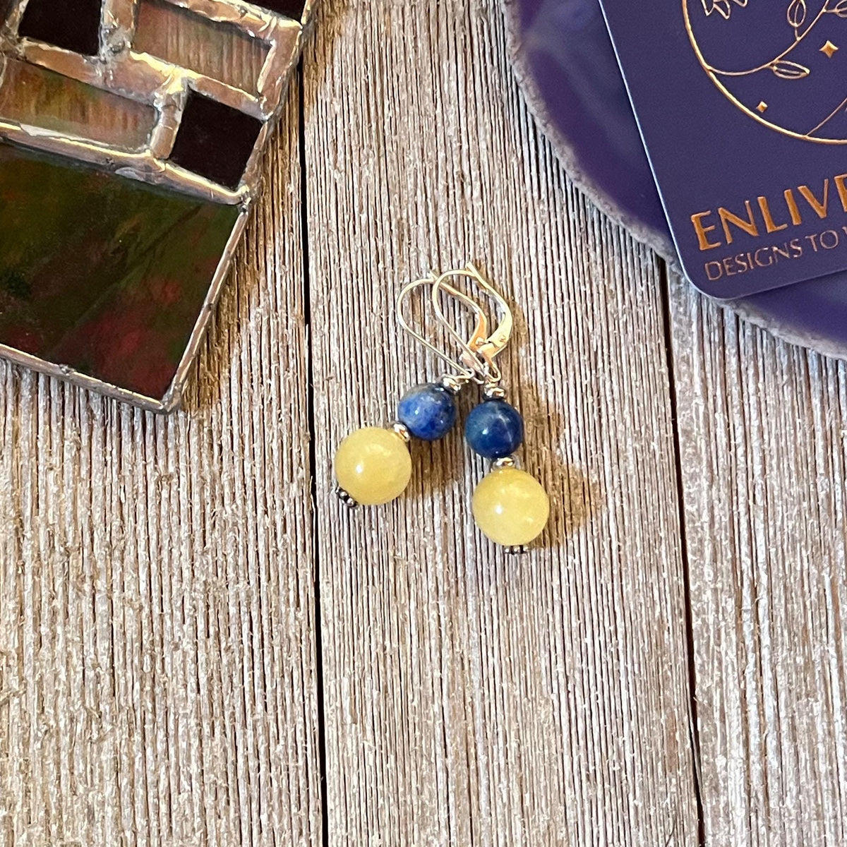Handmade Citrine Sodalite Spring Earrings: Geometric Circles Spheres, featuring yellow and blue beads, are shown on a wooden surface with a stained glass piece and an ENLIVEN DESIGNS TO card. Ideal for lovers of unique spring or geometric earrings.