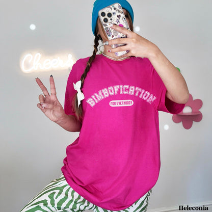 Woman in pink t-shirt with &amp;quot;BIMBOFICATION&amp;quot; text, holding phone, doing peace sign.
