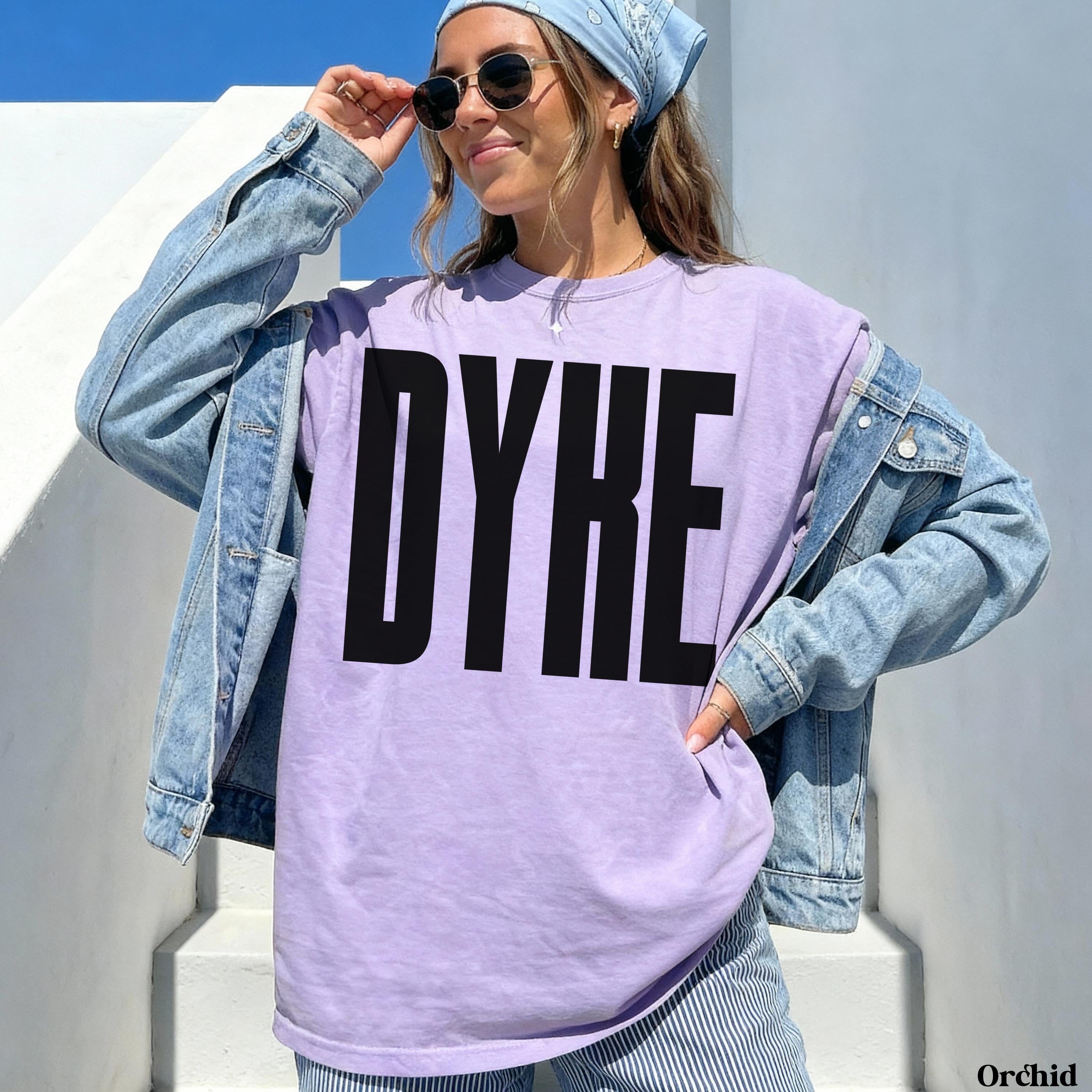 Woman in a denim jacket and bandana wearing a light purple t-shirt with &amp;quot;DYKE&amp;quot; in large black letters.