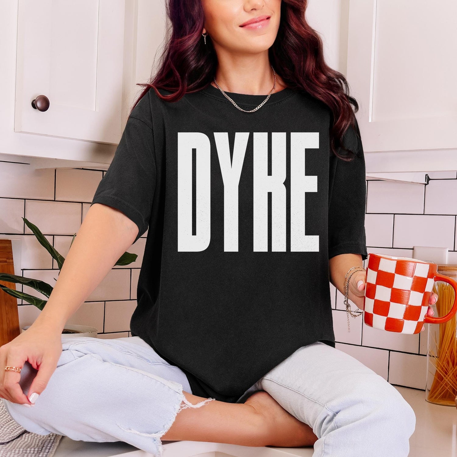 Black t-shirt with &amp;quot;DYKE&amp;quot; printed in white block letters, worn by a person holding a red and white checkered mug.