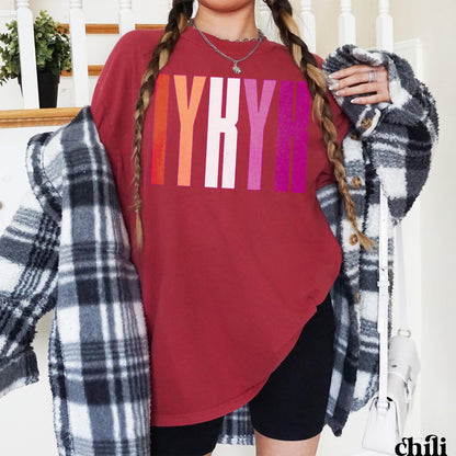 Woman wearing a maroon t-shirt with rainbow lettering and a plaid jacket.