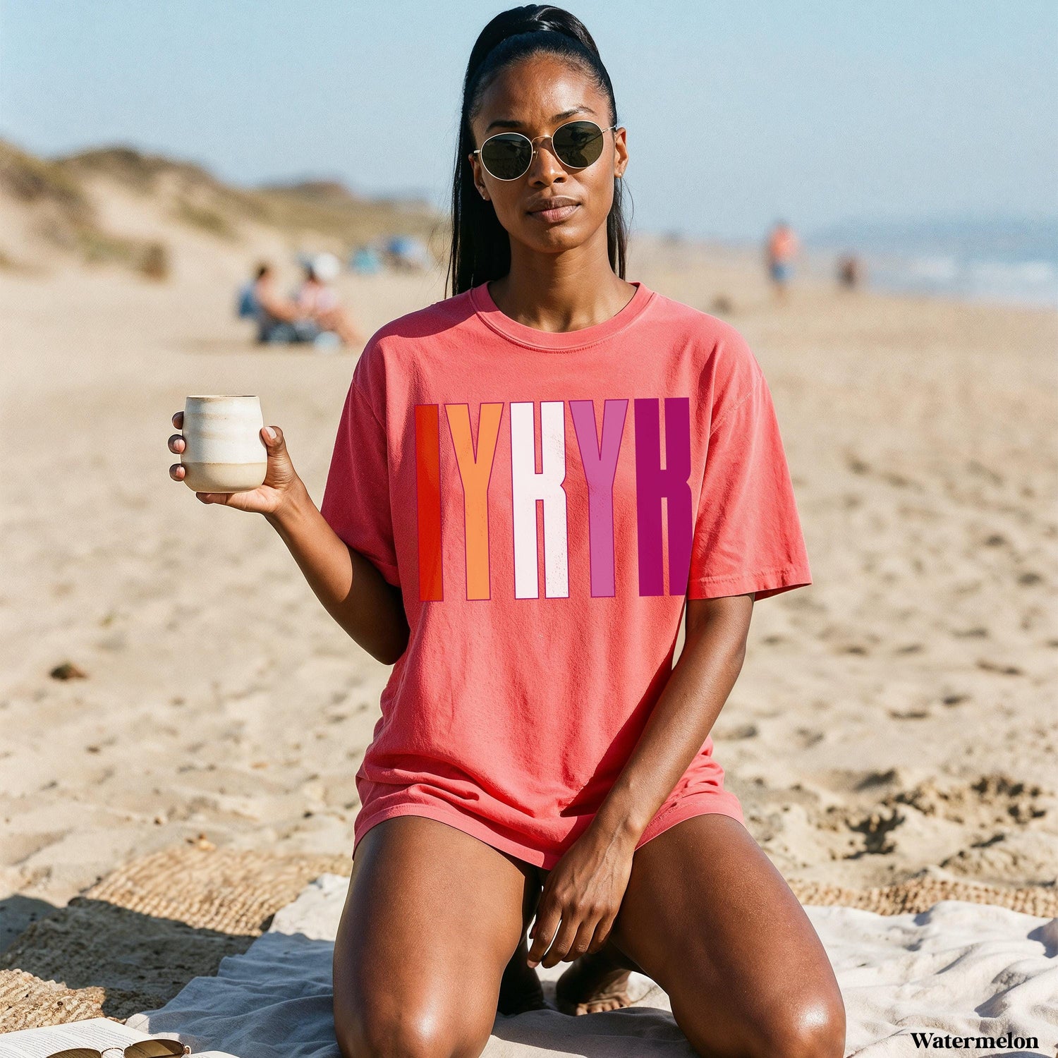 Woman in coral t-shirt with &amp;quot;IYKYK&amp;quot; in lesbian flag colors on a beach.