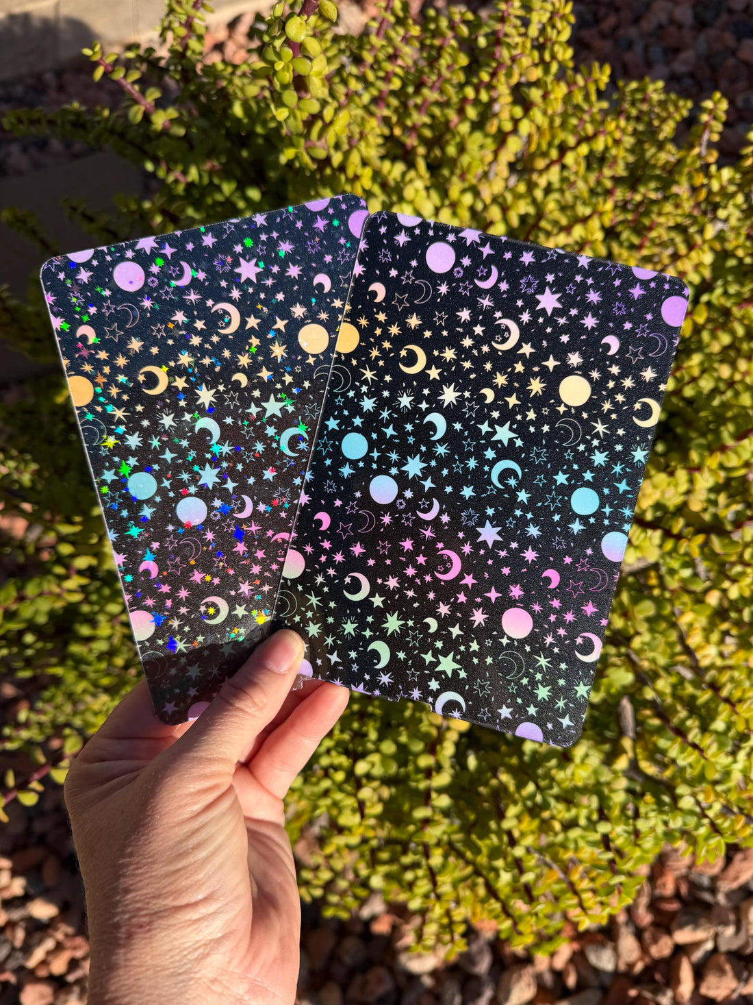 May include: Two rectangular cards with a celestial design. The cards feature a black background with holographic stars, moons, and pastel-colored circles. The cards are held up against a backdrop of green foliage.