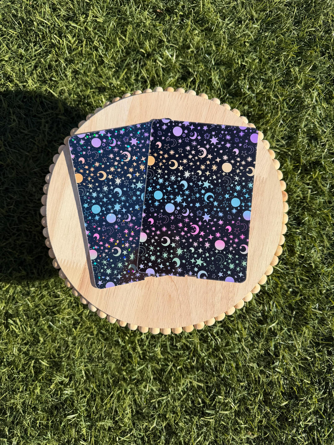 May include: Two notebooks with a black background and a celestial pattern of stars, moons, and planets in pastel colors. The notebooks are on a round wooden tray with a scalloped edge, set against a green grassy background.