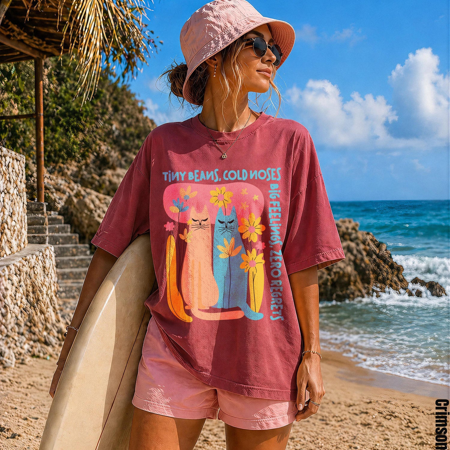 Woman in pink hat and sunglasses holding surfboard, wearing maroon t-shirt with colorful cat graphic.