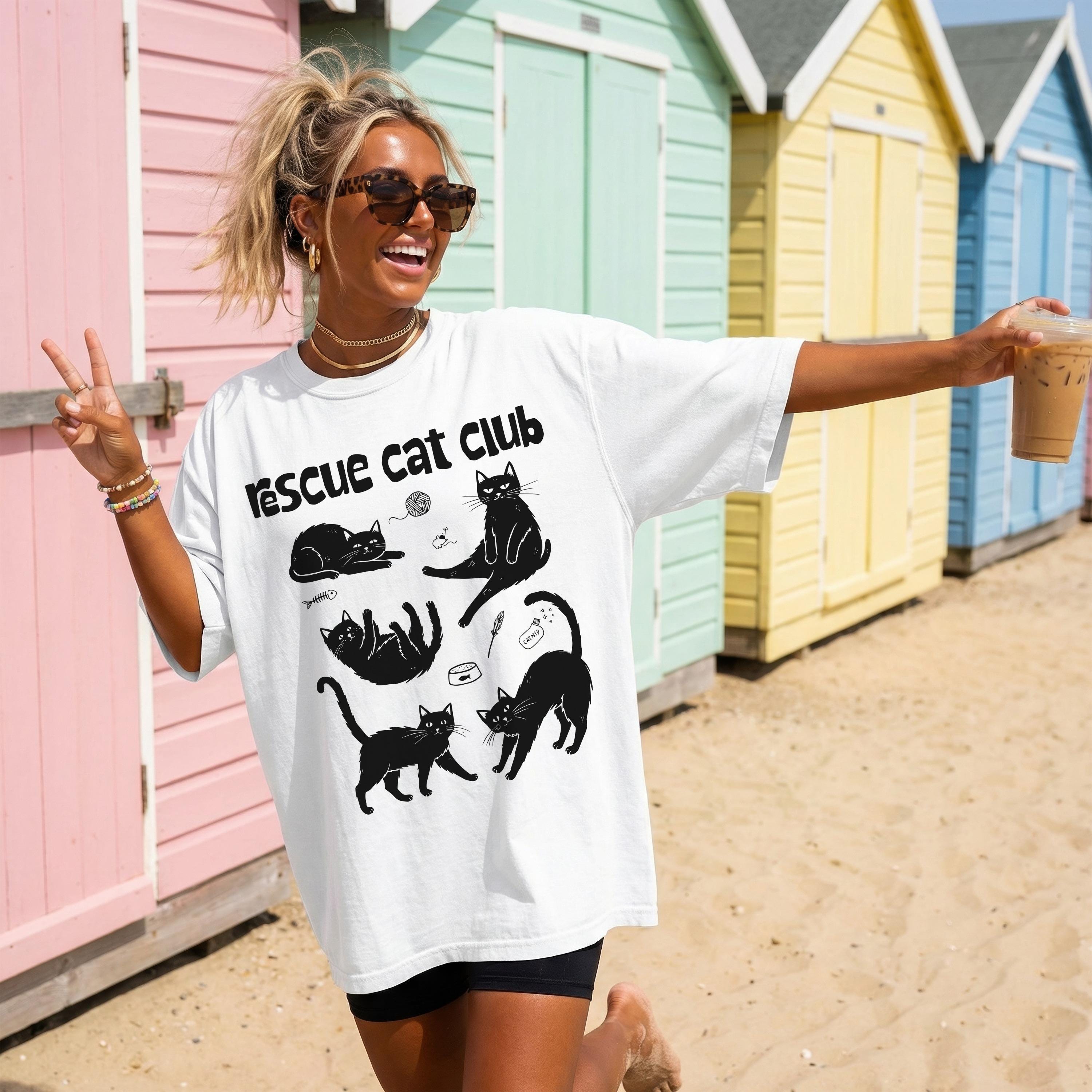 White t-shirt with &amp;quot;Rescue Cat Club&amp;quot; and cartoon cats, worn by a woman on a beach.