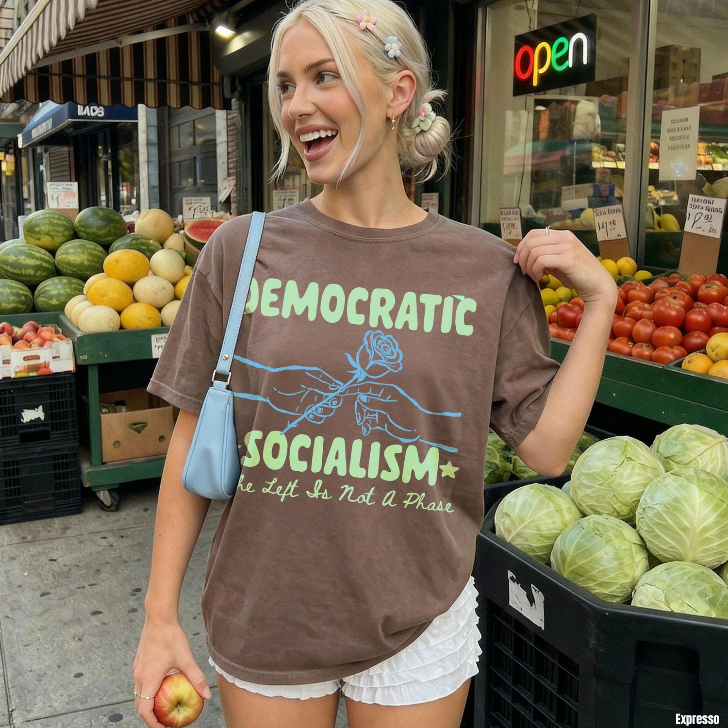 Woman in brown t-shirt with &amp;quot;Democratic Socialism&amp;quot; graphic, holding an apple outside a market.